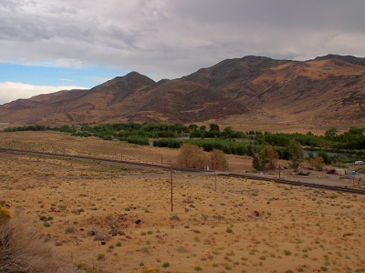 [Lots of barren-looking mountainy land except for the area near the river which is lined with trees. A railroad runs along a flat portion a short distance from the trees.]